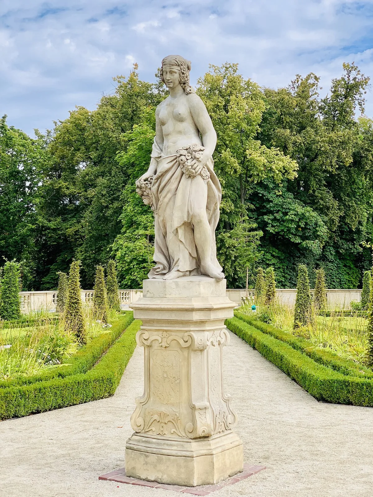 Stenen tuinbeeld van een vrouwelijke figuur op een sokkel tussen groene heggen in een formele tuin