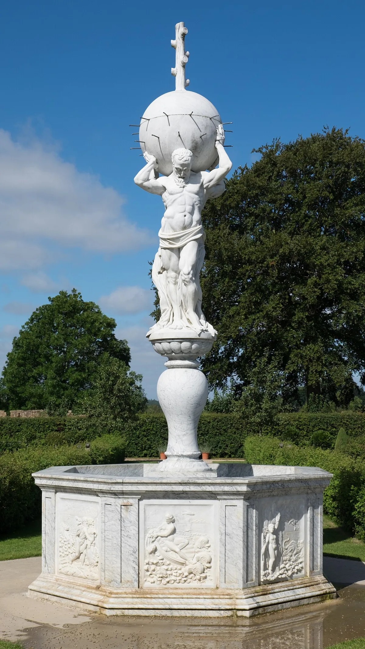 Marmeren tuinfontein met beeld van een figuur die een bol draagt, omgeven door groene struiken en bomen