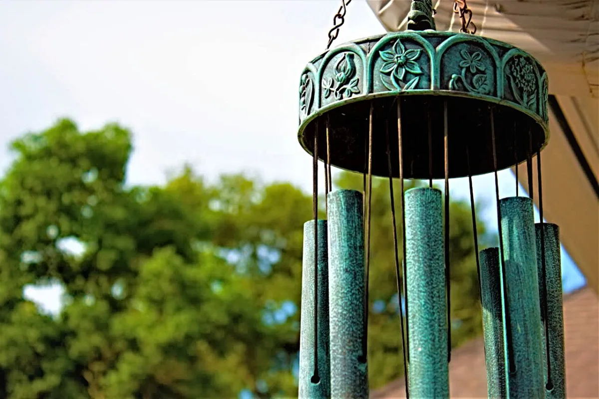 Windgong met groenpatina metalen buizen en bloemmotief, hangend voor een achtergrond van bomen
